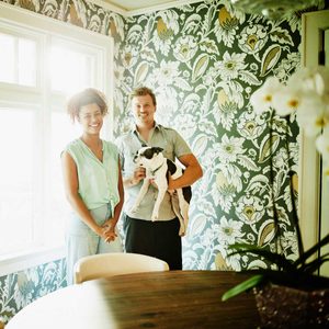 A couple stands smiling, holding a dog, in a brightly lit room with floral wallpaper and a round wooden table, surrounded by plants.