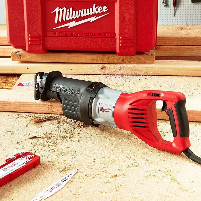 A Milwaukee reciprocating saw rests on a wooden work surface, surrounded by wood shavings, near a red tool case and replacement blades.