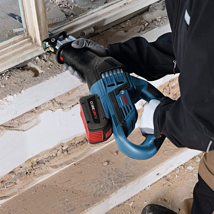 A power tool is being used to cut through a wooden frame in a construction area, with debris scattered on the floor and a partially dismantled wall visible.