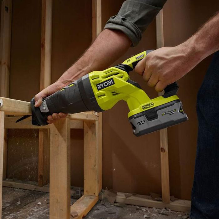 A reciprocating saw is being used to cut through a wooden beam in a construction setting, with debris scattering around in the background.
