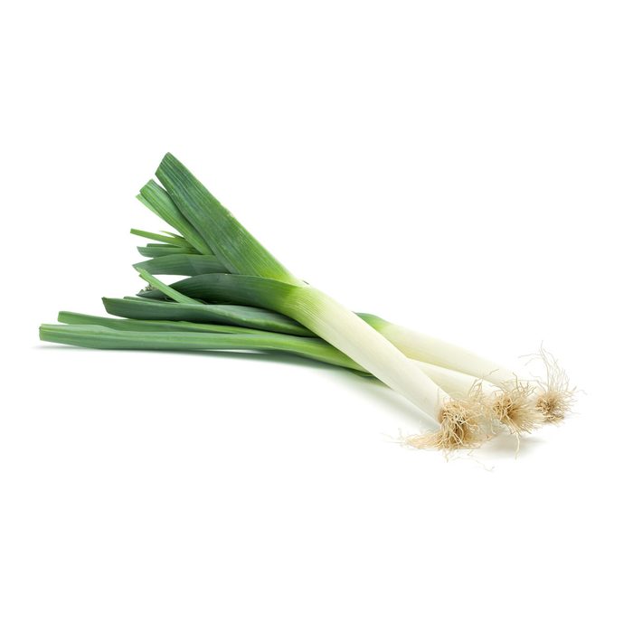 Three fresh leeks lie on a white surface, showcasing their long green tops and smooth white bottoms with roots extending from the ends.