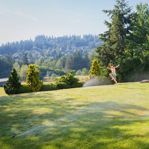 A child runs joyfully through green grass, splashing water from nearby sprinklers under a bright sky, surrounded by trees and distant hills.