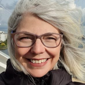 A woman with silver hair and glasses smiles, her hair blowing in the wind; she's outdoors near water and buildings under a cloudy sky.