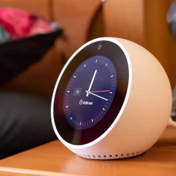 A round, white smart clock displays the time at 6:00 am on a wooden surface, surrounded by a blurred background of furniture and colorful bedding.