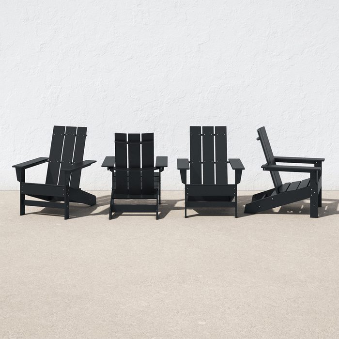 Four black Adirondack chairs are positioned next to each other on a light-colored concrete surface against a plain white wall.