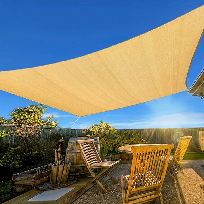 A large beige shade sail hangs above a wooden patio, providing shelter over a round table and several folding chairs, with greenery in the background.