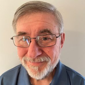 A middle-aged man smiles, wearing glasses and a blue shirt, against a light gray wall that provides a neutral background.