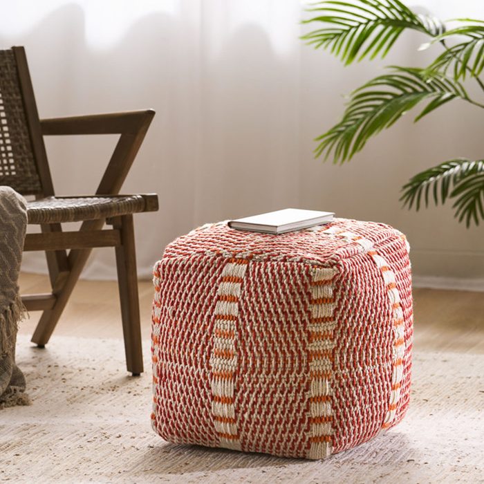 A colorful woven pouf sits on a light rug, supporting a small book, near a wooden chair and a potted plant in a bright room.