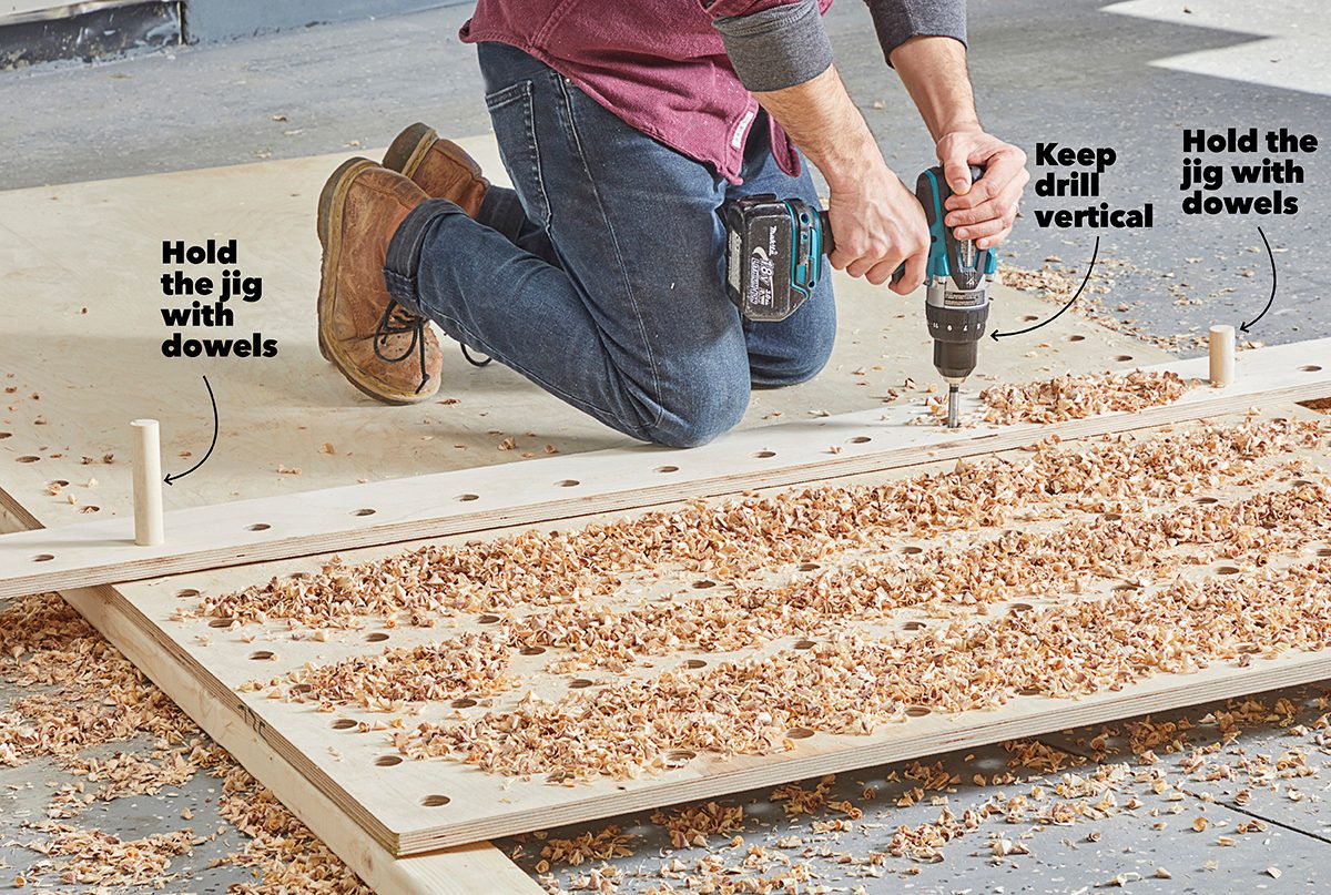 A person drills into a wooden board placed on the floor, surrounded by wood shavings, while holding a jig with dowels for stability.