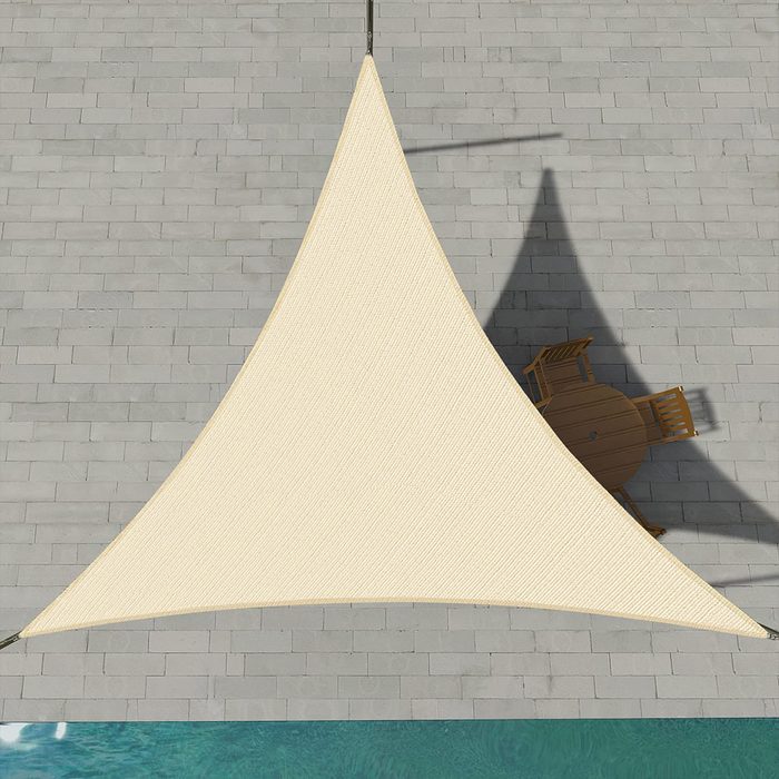 A large triangular shade sail is suspended above a patio, casting a shadow over a round table and chairs adjacent to a swimming pool.