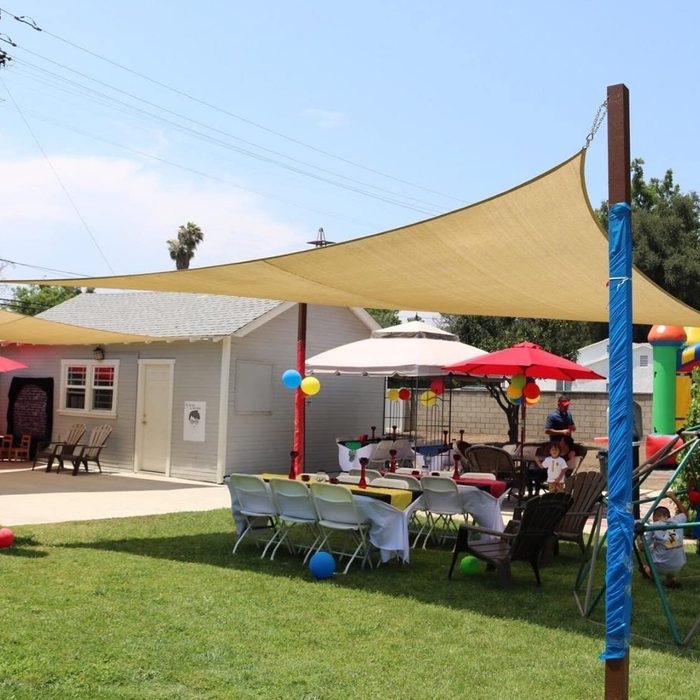 A large shade sail provides cover over tables and chairs set up in a vibrant outdoor party space, surrounded by decorations and play structures.
