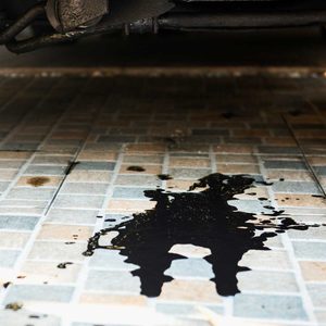 A dark liquid spills onto a patterned tiled surface, pooling and spreading beneath a vehicle in a partially shaded garage environment.