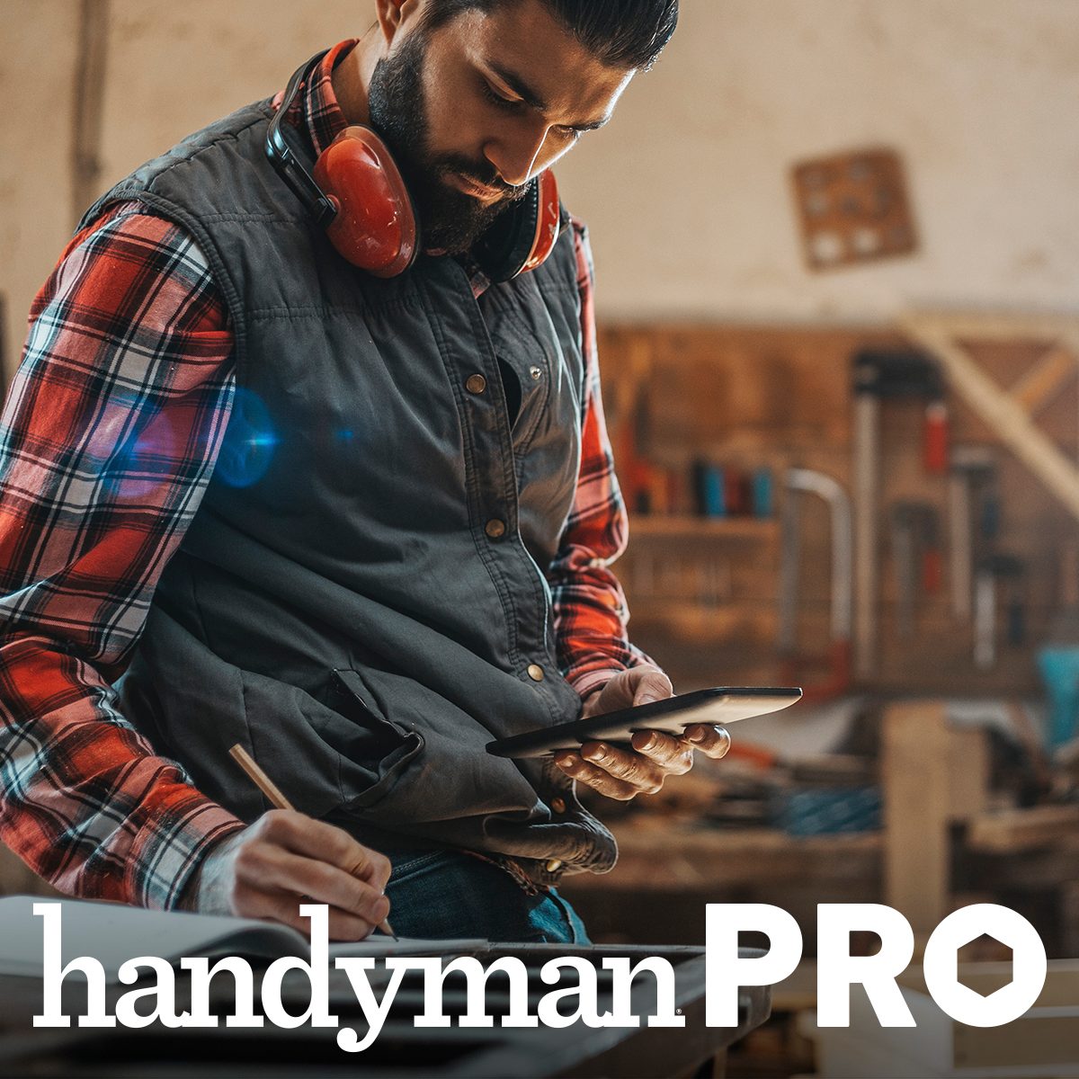 A man wearing safety headphones reviews plans on a tablet while seated at a workbench in a workshop filled with tools and woodwork materials.