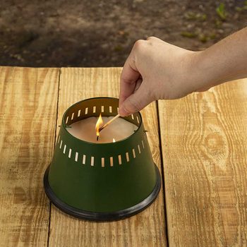 A hand holds a match, igniting a candle inside a green container. The scene is set on a wooden table in a natural outdoor environment.
