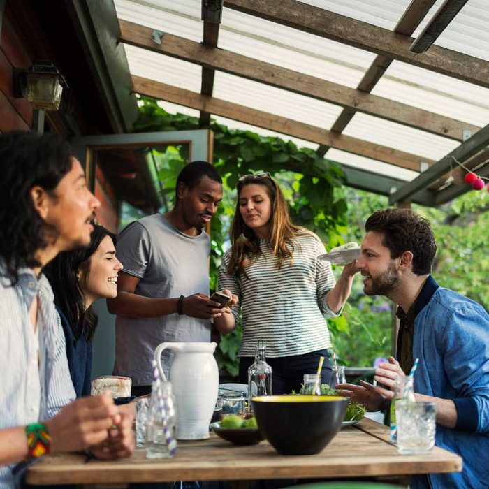 A group of five friends engages in conversation around a table filled with food and drinks on a covered outdoor patio surrounded by greenery.