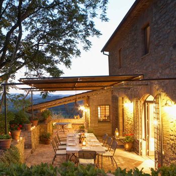 A long dining table is set outside, illuminated by soft lights, next to a rustic stone house surrounded by greenery and a scenic view at dusk.