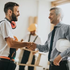 A construction worker shakes hands with a businessman in a well-lit room, while a colleague in the background manages a ladder, suggesting a work-related discussion.