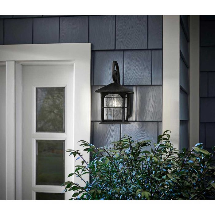 A black lantern hangs beside a white door, glowing softly, casting light on the textured blue wall and nearby green foliage.