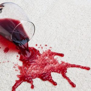 A wine glass lies overturned, spilling red liquid onto a light-colored carpet, creating a pool and splatter effect in the surroundings.