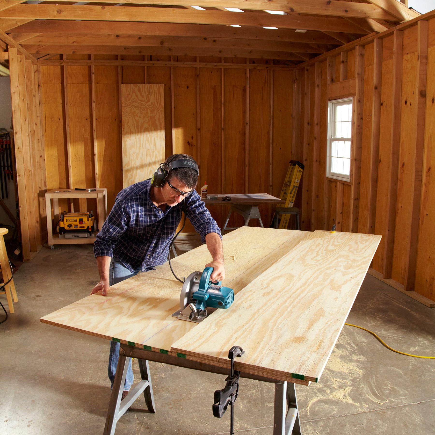 A man uses a circular saw on a large piece of wood, working in a wooden workshop with light spilling across the floor and tools visible.