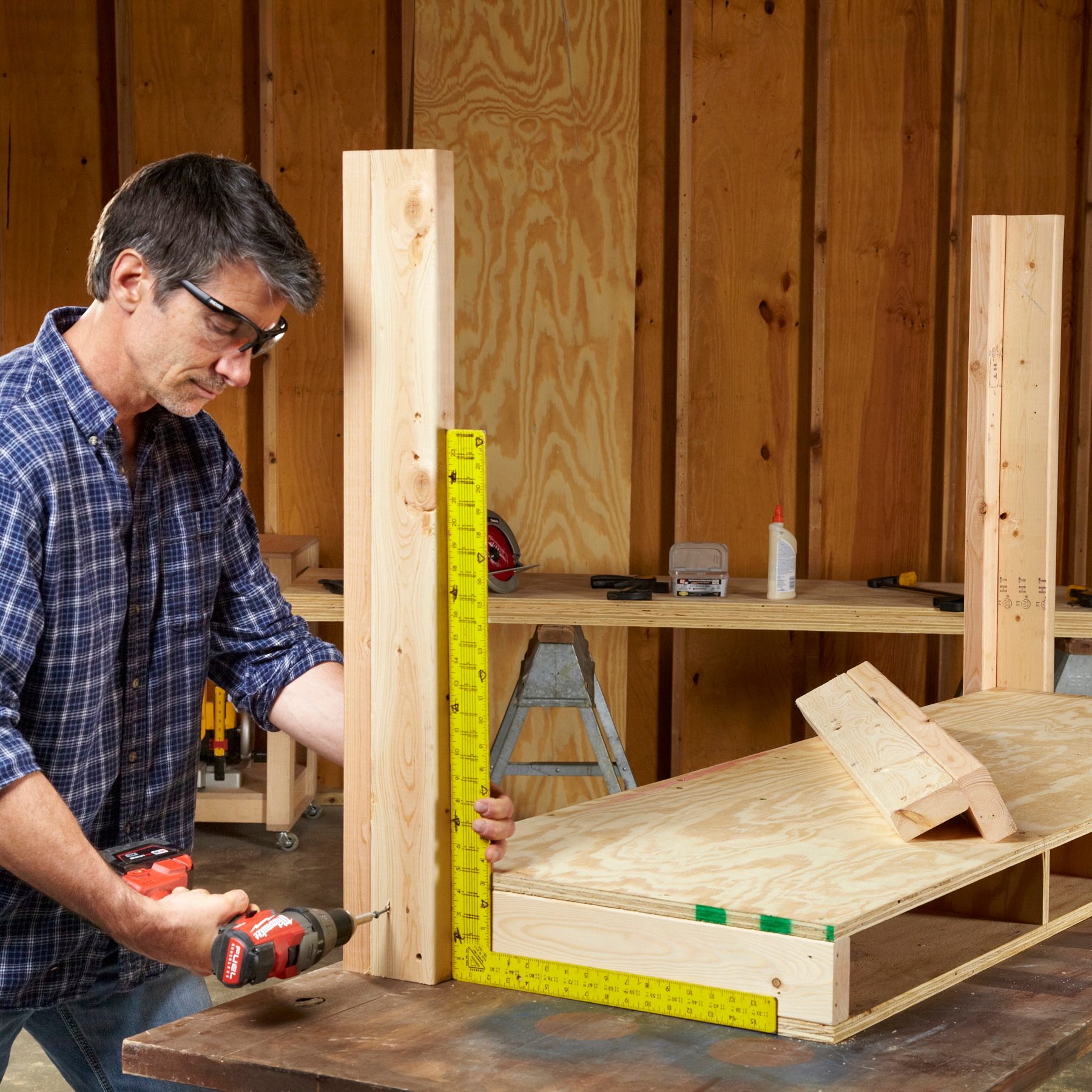 A man drills into a wooden frame using a measuring square, surrounded by tools and wood in a workshop with wooden walls.
