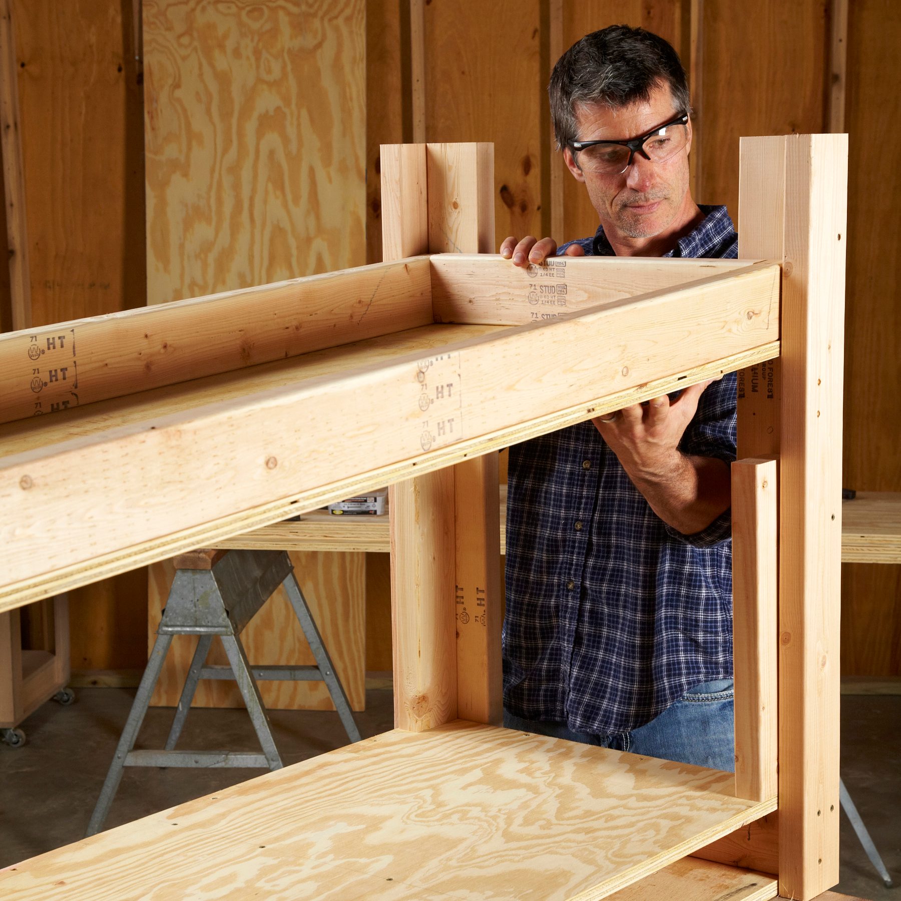 A man assembles wooden shelving, carefully positioning a shelf between two vertical supports in a workshop with exposed wood walls and tools nearby.