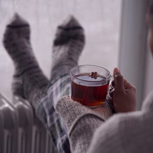 A person holds a transparent cup of tea while resting their feet on a radiator, wearing cozy socks and a chunky sweater.