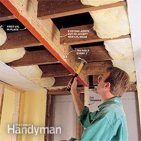A person hammers a nail into a wooden beam, surrounded by exposed ceiling joists and insulation in a construction environment.