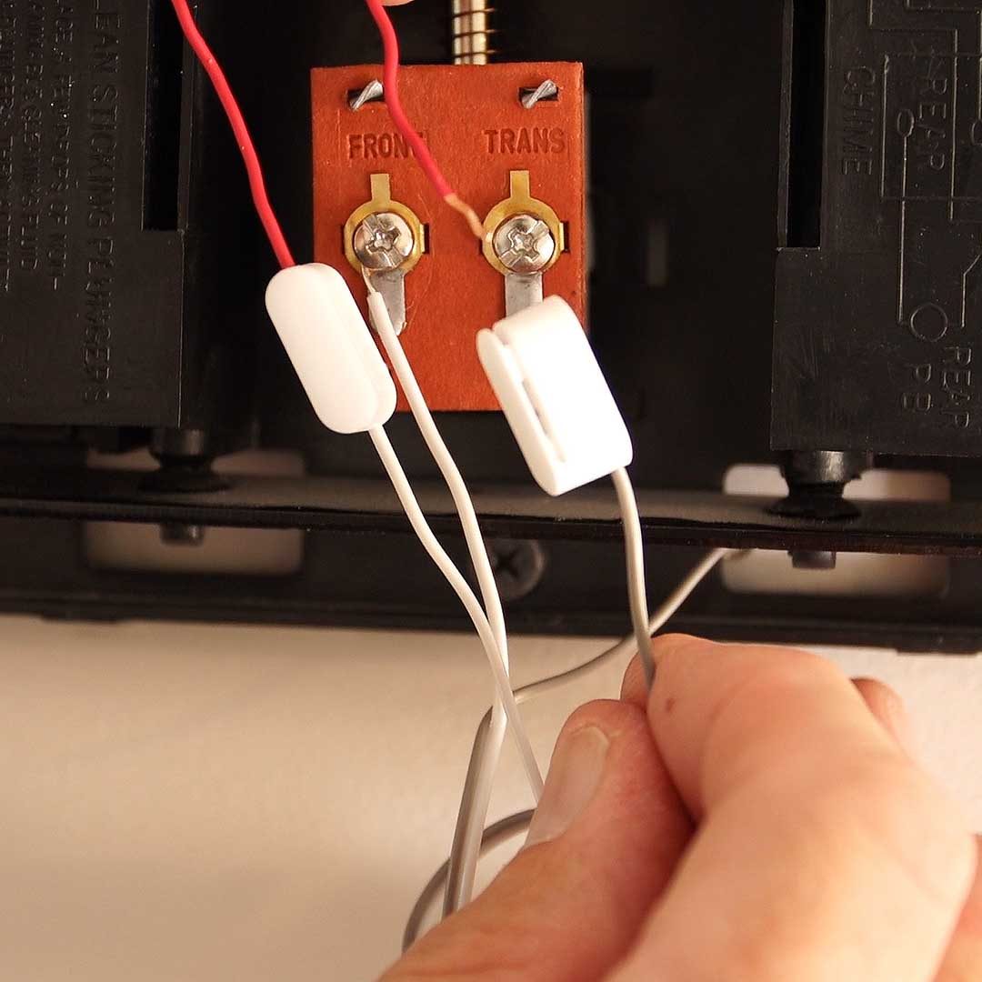A hand connects wires to terminals on a brown plate, with a black device in the background and a white wall as the context.
