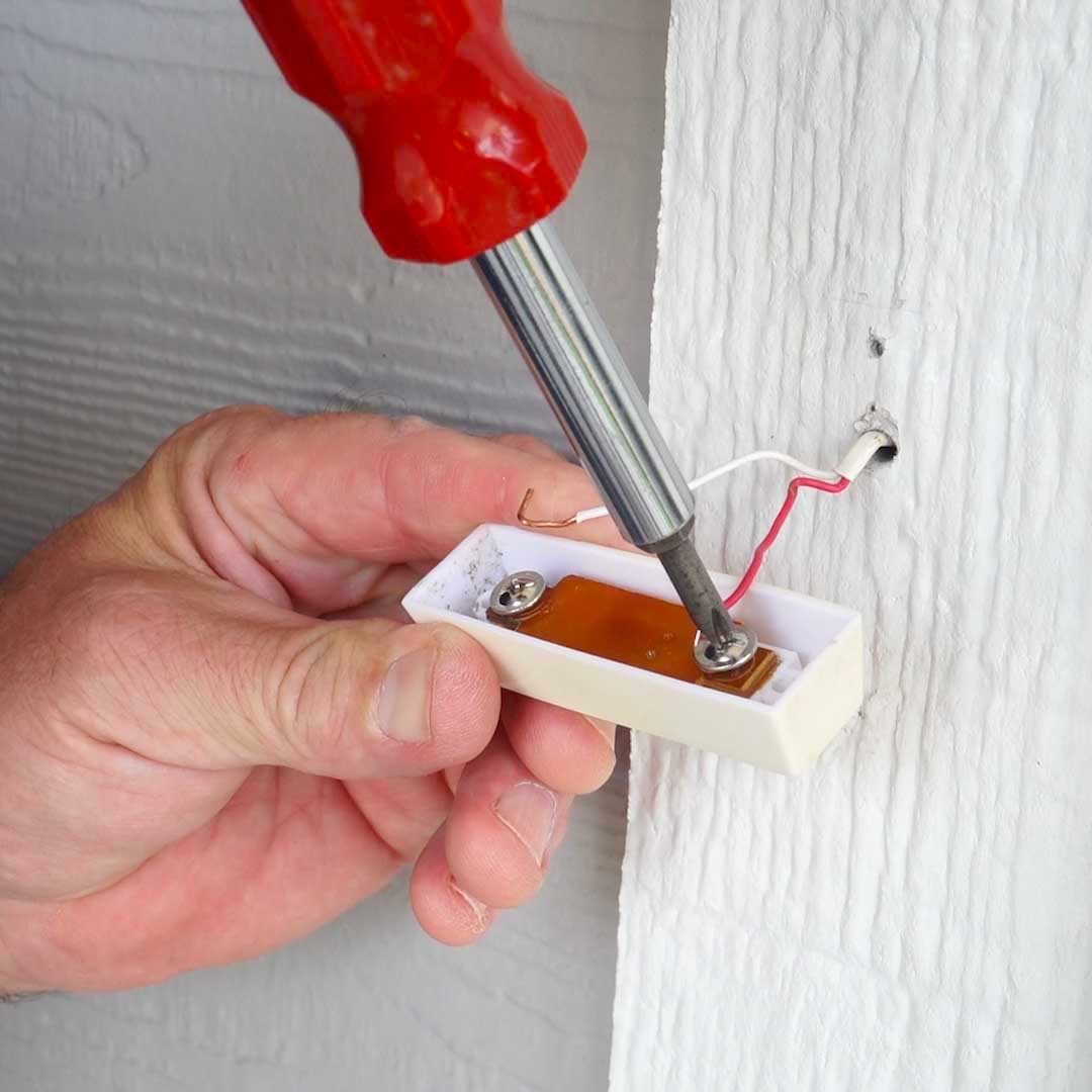 A hand holds a screwdriver, fastening a small white battery holder against a textured wall, with wires connected nearby.