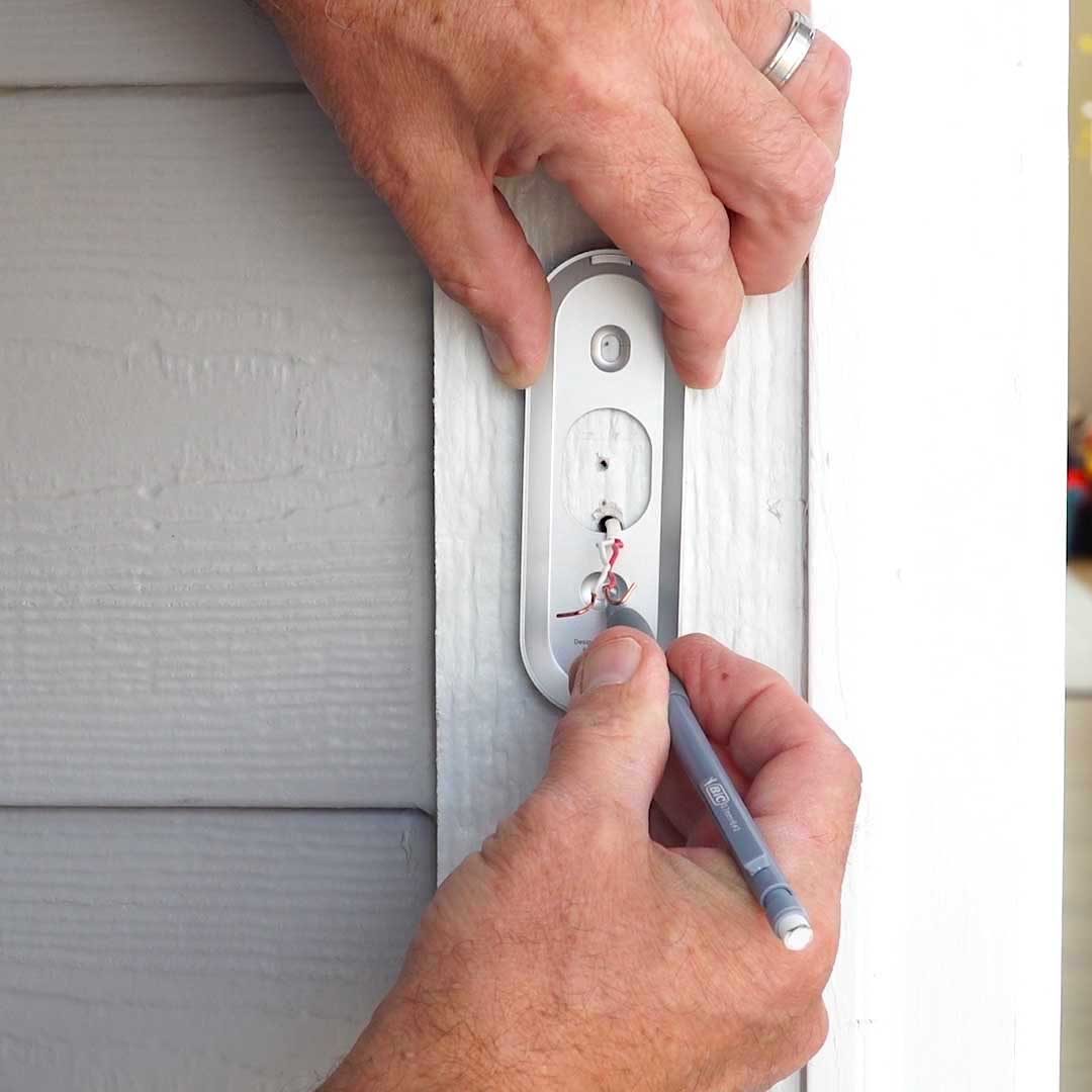 A hand holds a pen, making adjustments to a lock installation on a door, with a light gray wooden surface surrounding it.
