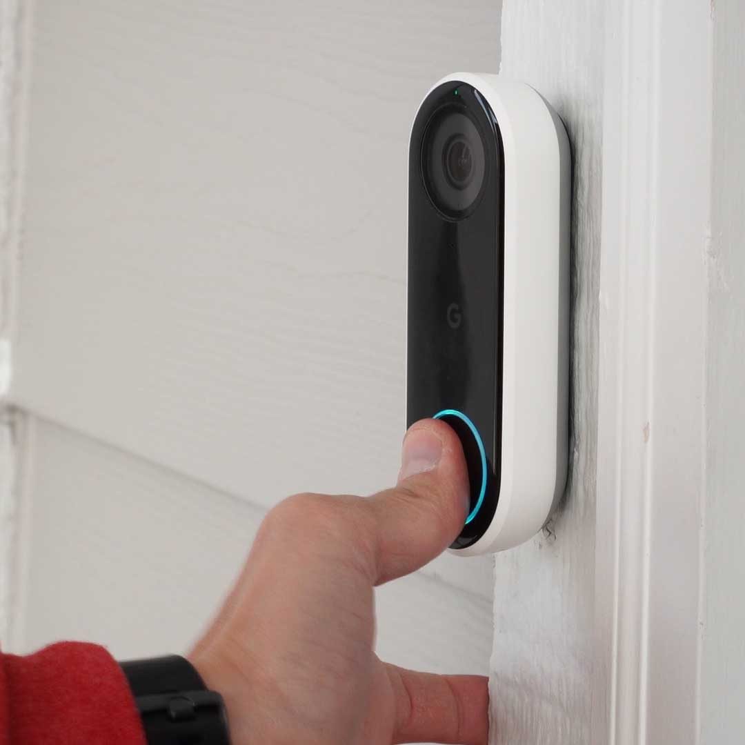 A hand presses a doorbell camera mounted on a wall, which is designed to monitor entryways. The surrounding surface is light-colored wood.