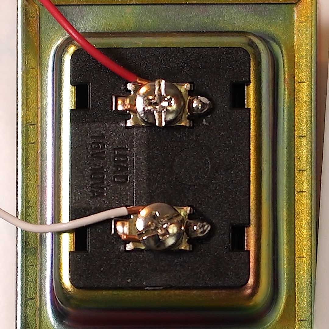 Two metallic connectors are soldered to wires, positioned on a black base. The surrounding area is metallic with some reflective surfaces.