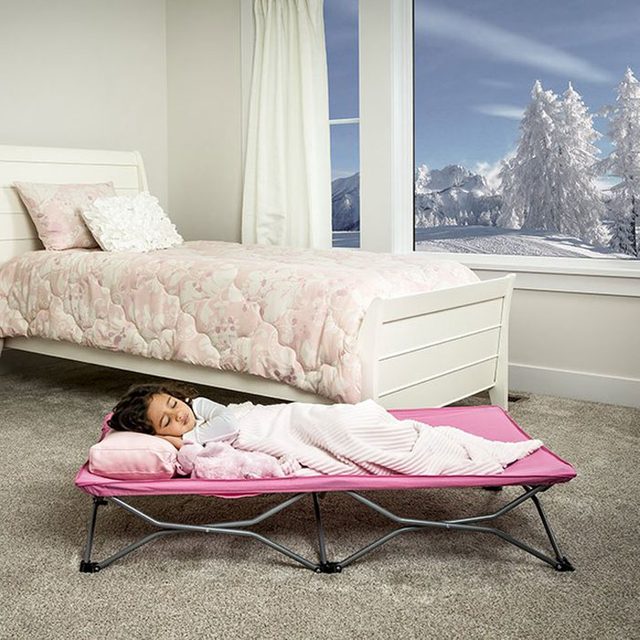 A girl sleeps on a pink cot in a cozy room, beside a bed, with snowy mountains visible through a window.
