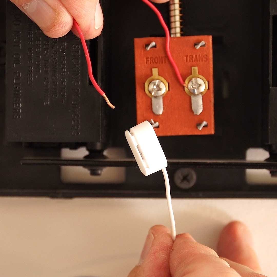 A hand holds a red wire above a white connector, positioned near a circuit board with metal terminals, integrated into a device on a white surface.