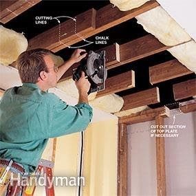A man uses a circular saw to cut wooden beams above him while standing on a ladder in a partially finished interior space.
