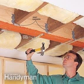 A man uses a hammer to secure joist hangers and a steel strap in a ceiling, surrounded by exposed wooden beams and insulation.