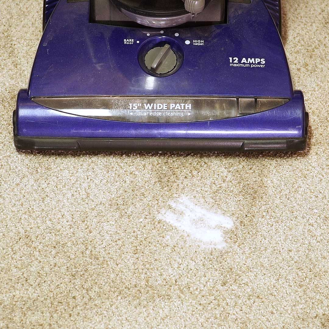 A vacuum cleaner is positioned on a light-colored carpet, actively cleaning and revealing a clear patch of lighter carpet beneath it.