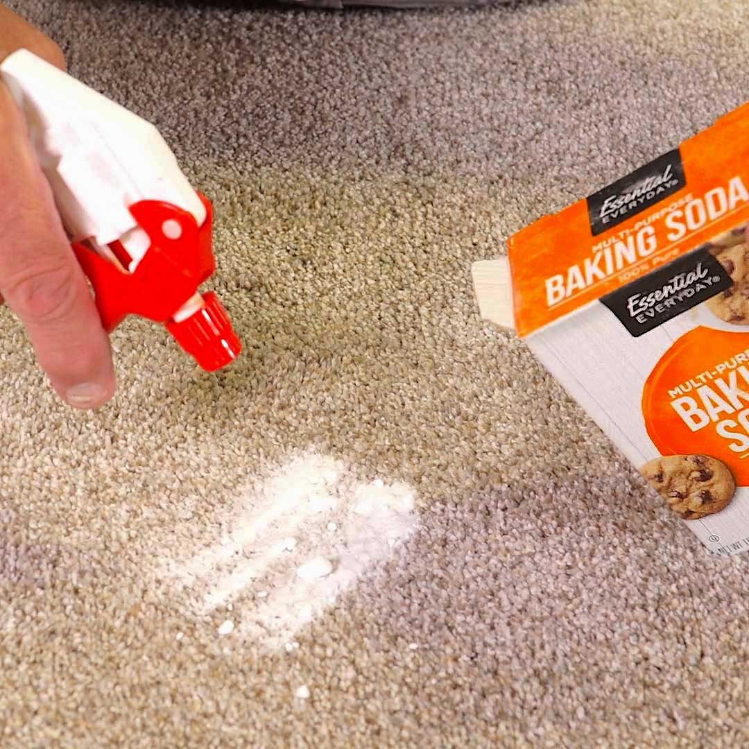A hand with a spray bottle applies a mist over a patch of baking soda on textured carpet, indicating a cleaning process in progress.