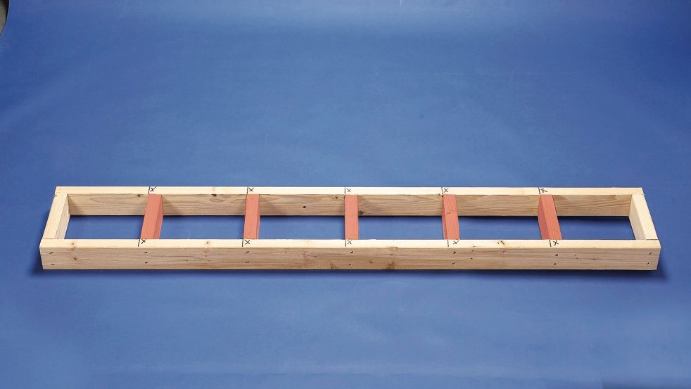 A wooden frame with spaced red sections lies flat on a blue surface, likely designed for alignment or measurement tasks in a workshop setting.