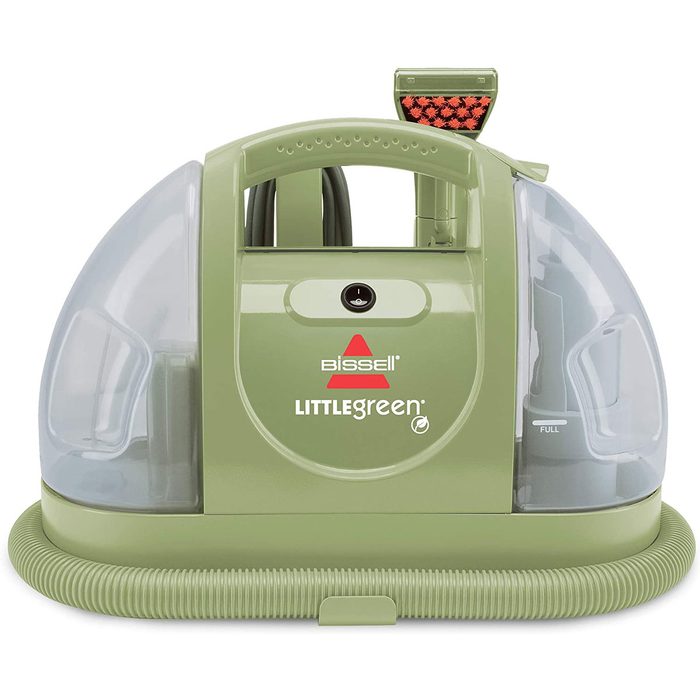 A small, green carpet cleaner sits upright, featuring transparent water tanks and a hose attachment, designed for cleaning upholstery and carpets efficiently.