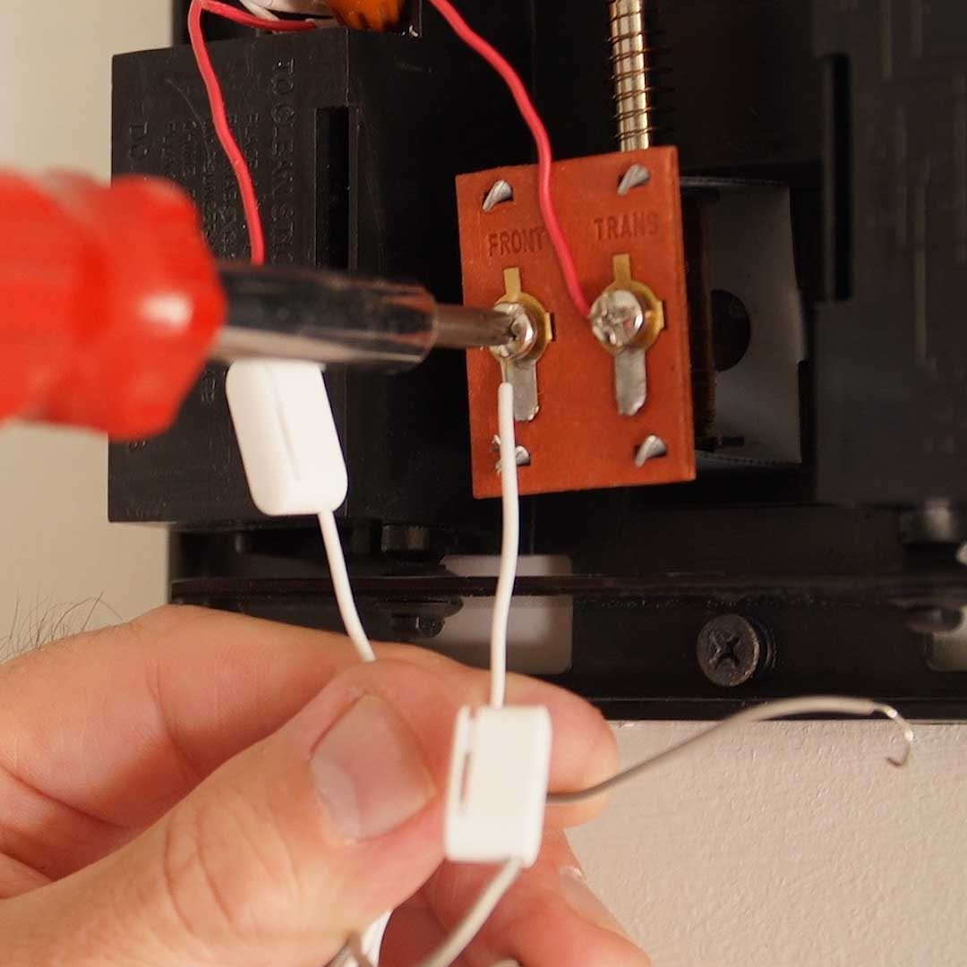 A hand uses a screwdriver to tighten wires into a terminal block labeled "FRONT" and "TRANS," mounted on a black electrical device on a wall.
