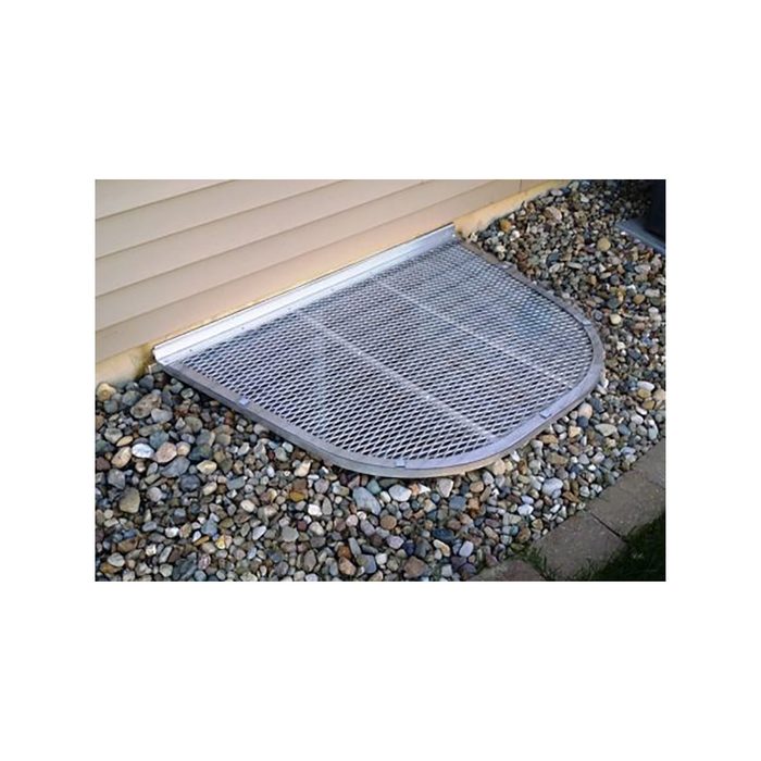 A metal grating covers a circular area at the base of a house, surrounded by small stones and grass, providing ventilation or access to a basement.