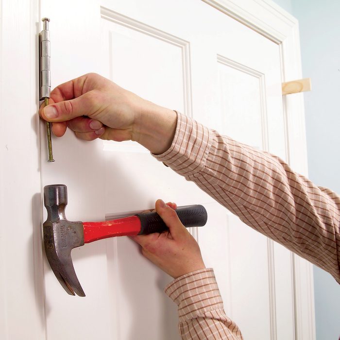 A person is installing a door hinge, using a hammer to drive in a screw on a white door, set against a light-colored wall.