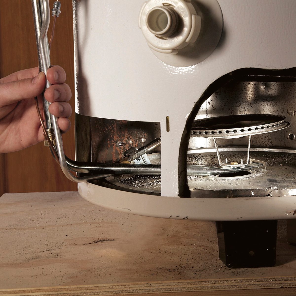 A hand adjusts a gas pipe connected to a white appliance, revealing internal components on a wooden surface with some debris.