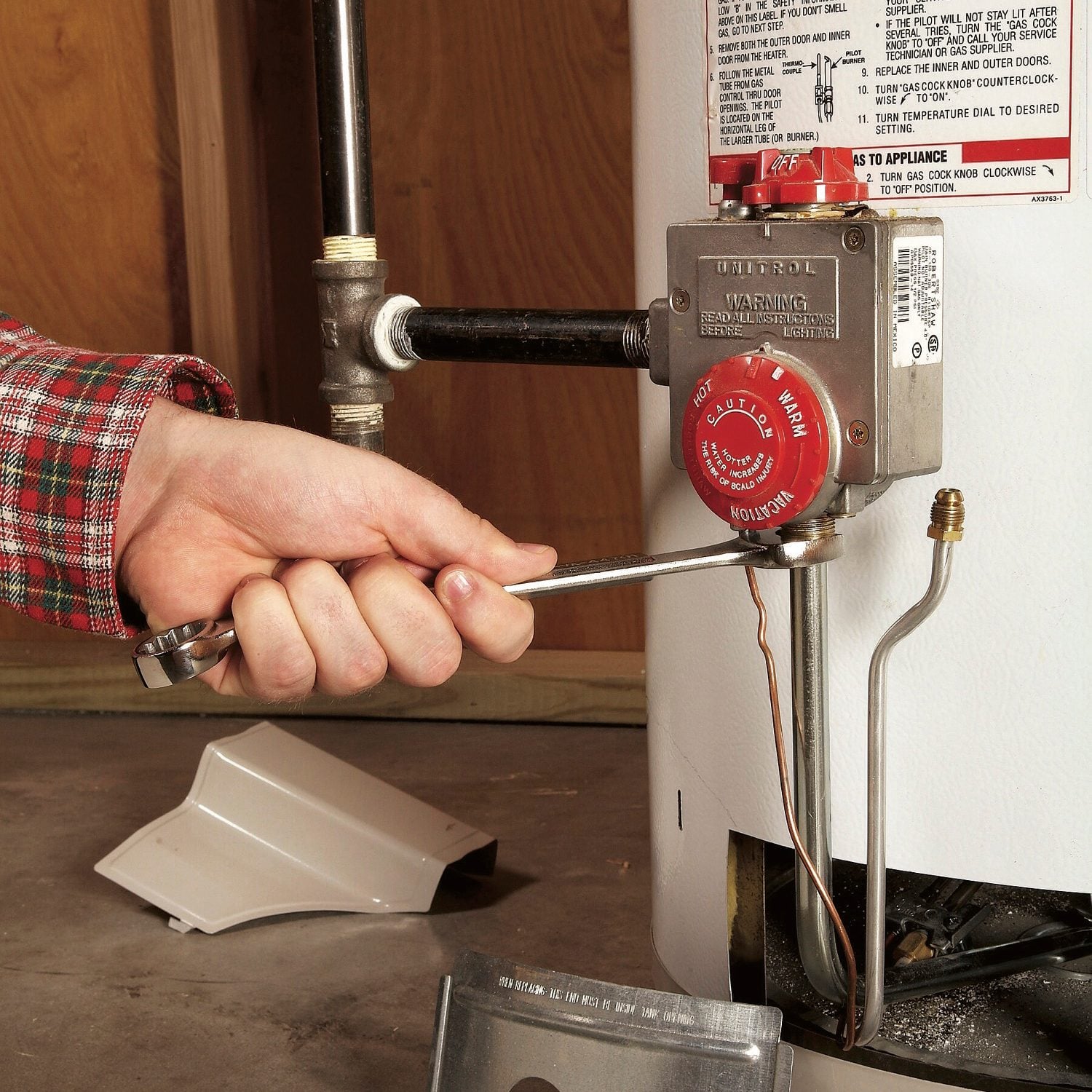 A person uses a wrench to tighten a gas control valve on a water heater, positioned on a concrete floor with wooden walls in the background.