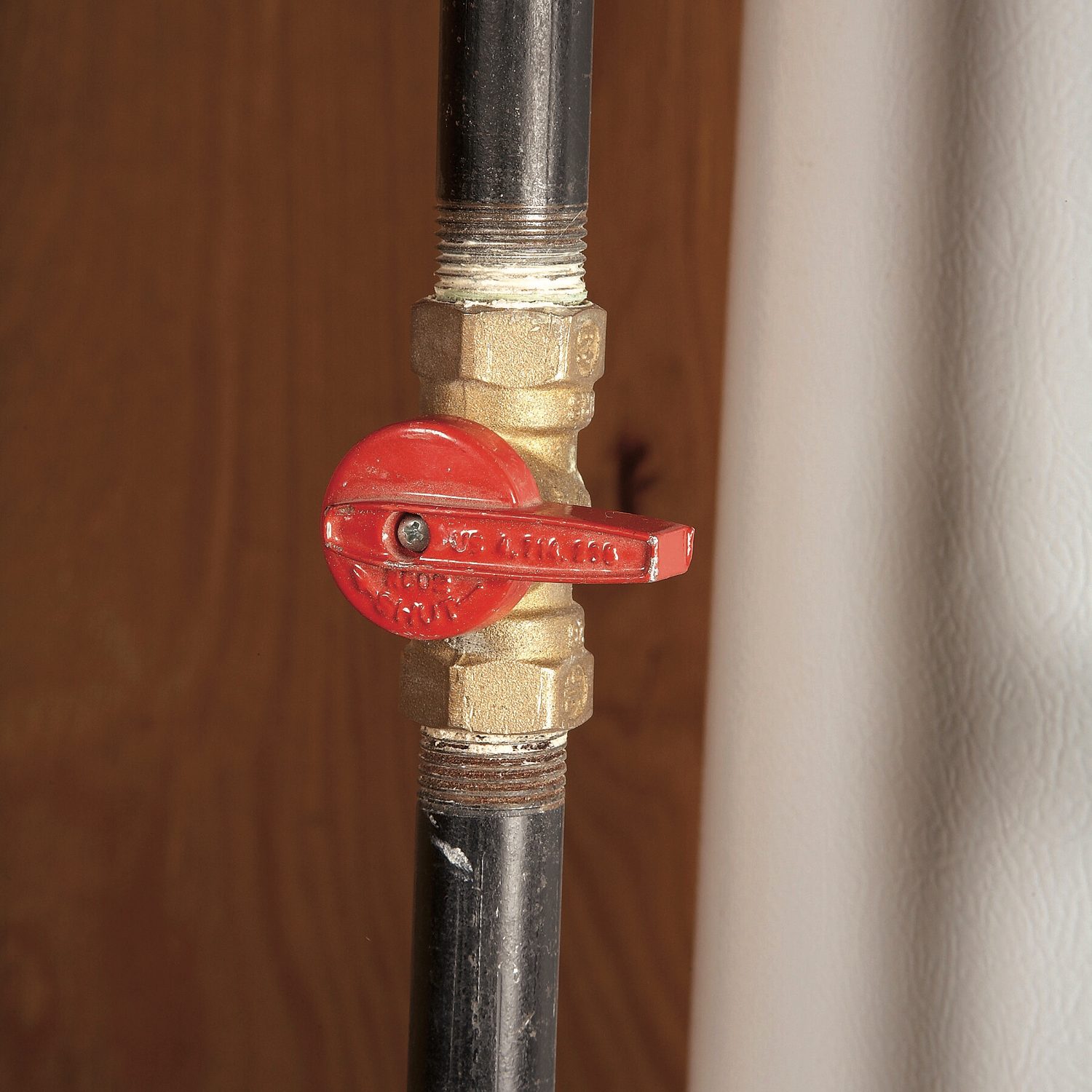 A brass valve with a red handle is positioned on a black pipe, set against a brown wooden background. The valve's handle suggests it can control flow.