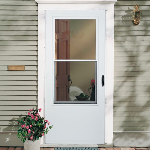 A white screen door stands closed, framed by beige siding. A potted flower arrangement is placed alongside, with a faint reflection of a room visible through the glass.
