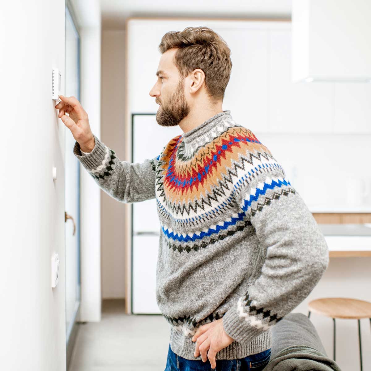 A man adjusts a wall thermostat while standing in a modern kitchen, wearing a colorful, patterned sweater, with minimalistic furnishings visible in the background.