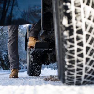 A person is stepping out of a vehicle onto a snowy road, with a close-up view of a tire and winter scenery in the background.